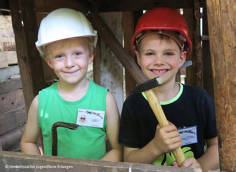 News-Bild Baustelle: Zwei Kinder in einer Holzhütte mit Bauhelmen auf dem Kopf und Werkzeug in der Hand